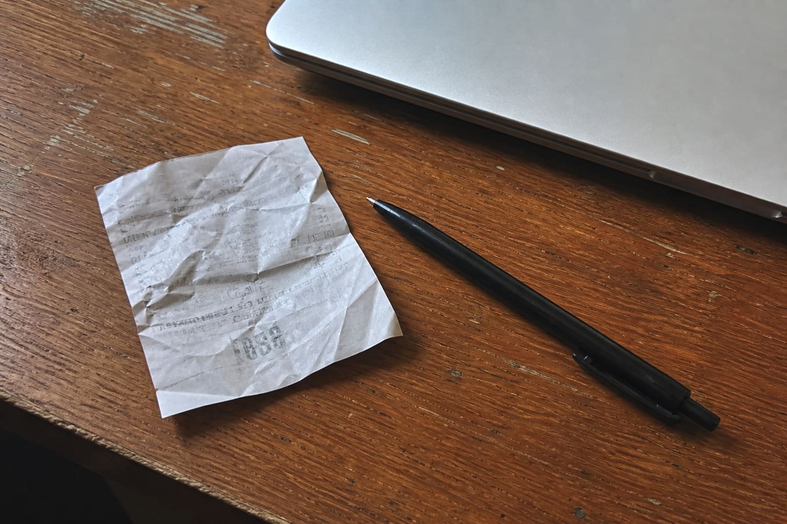 Receipt and pen on a wooden desk next to a laptop